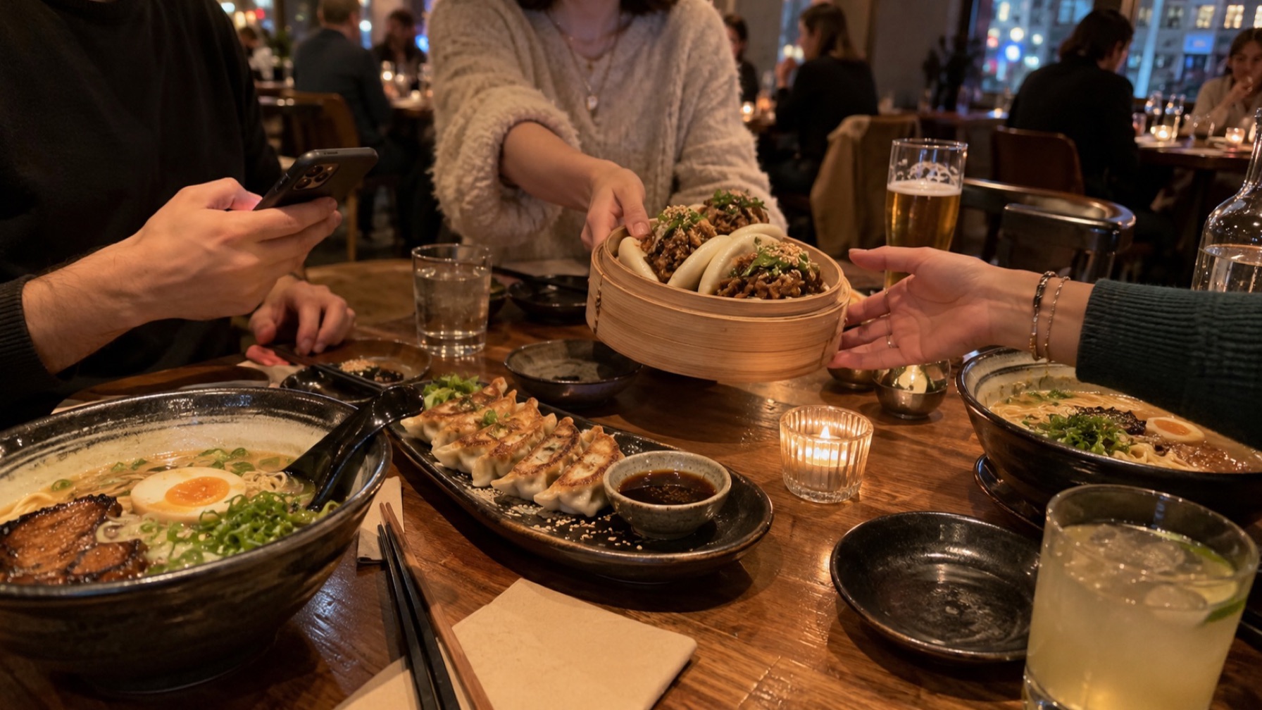 Troje znajomych dzieli się daniami przy stole w restauracji: bao, ramen i gyoza; ktoś trzyma telefon nad stołem.